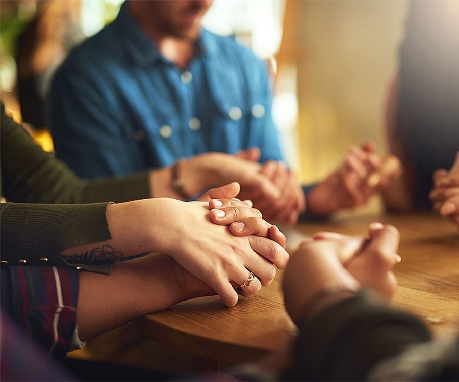 Friends and family sit in circle holding hands around a table.